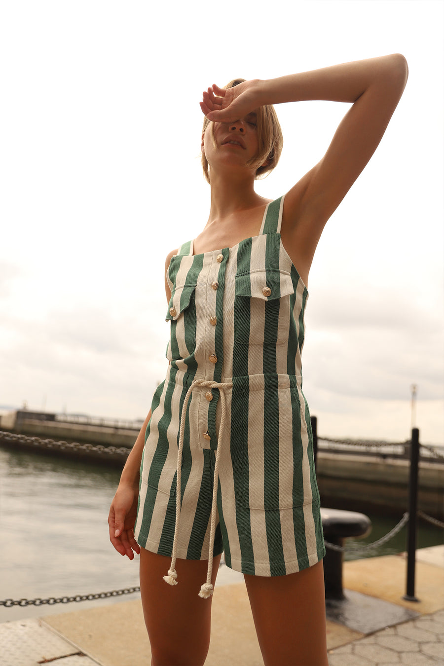 This is a photo of a woman wearing a green striped romper with a rope belt at the waist and gold buttons down the center front. She stands in front of a body of water.
