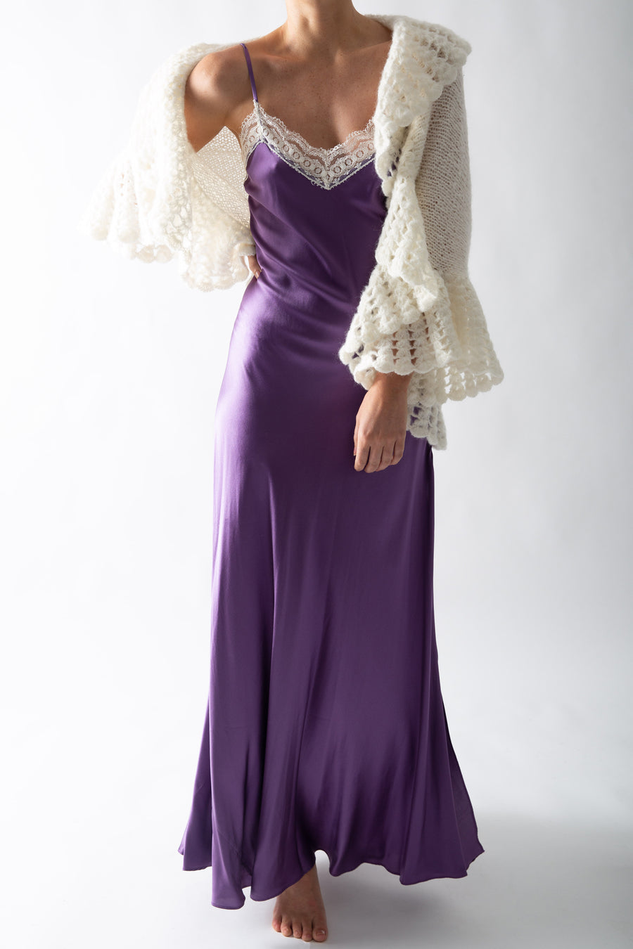 This is a photo of a woman wearing a plum colored silk slip dress with lace trim at the neckline. She wears a white knit ruffle cardigan overtop.