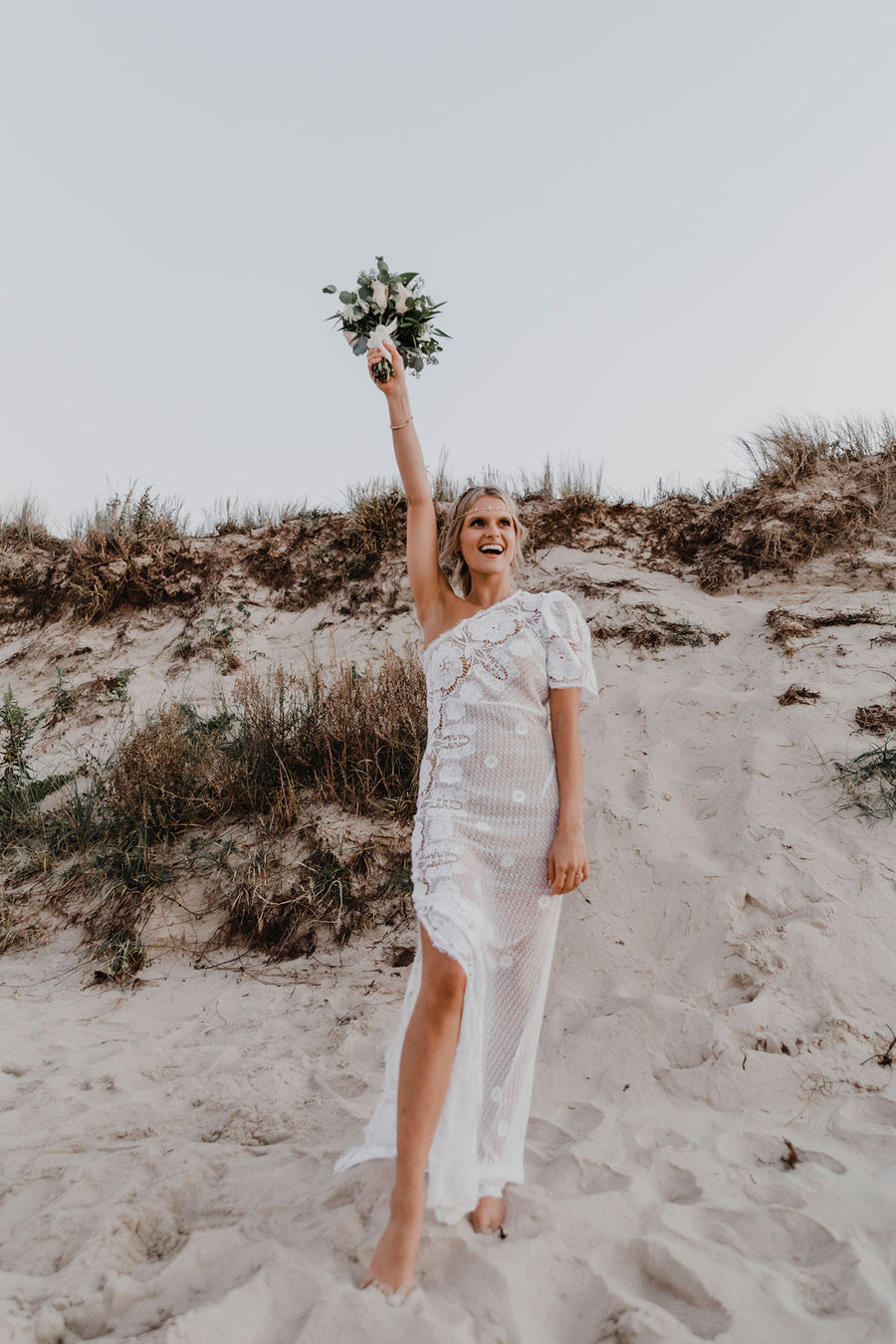 This is a photo of a bride wearing a floor length, one shoulder, maxi lace dress. There is a scalloped leg slit on the same side as the strapless top part of dress. The dress is lined to the leg slit. She smiles as she stands on a beach holding her bouquet of flowers.