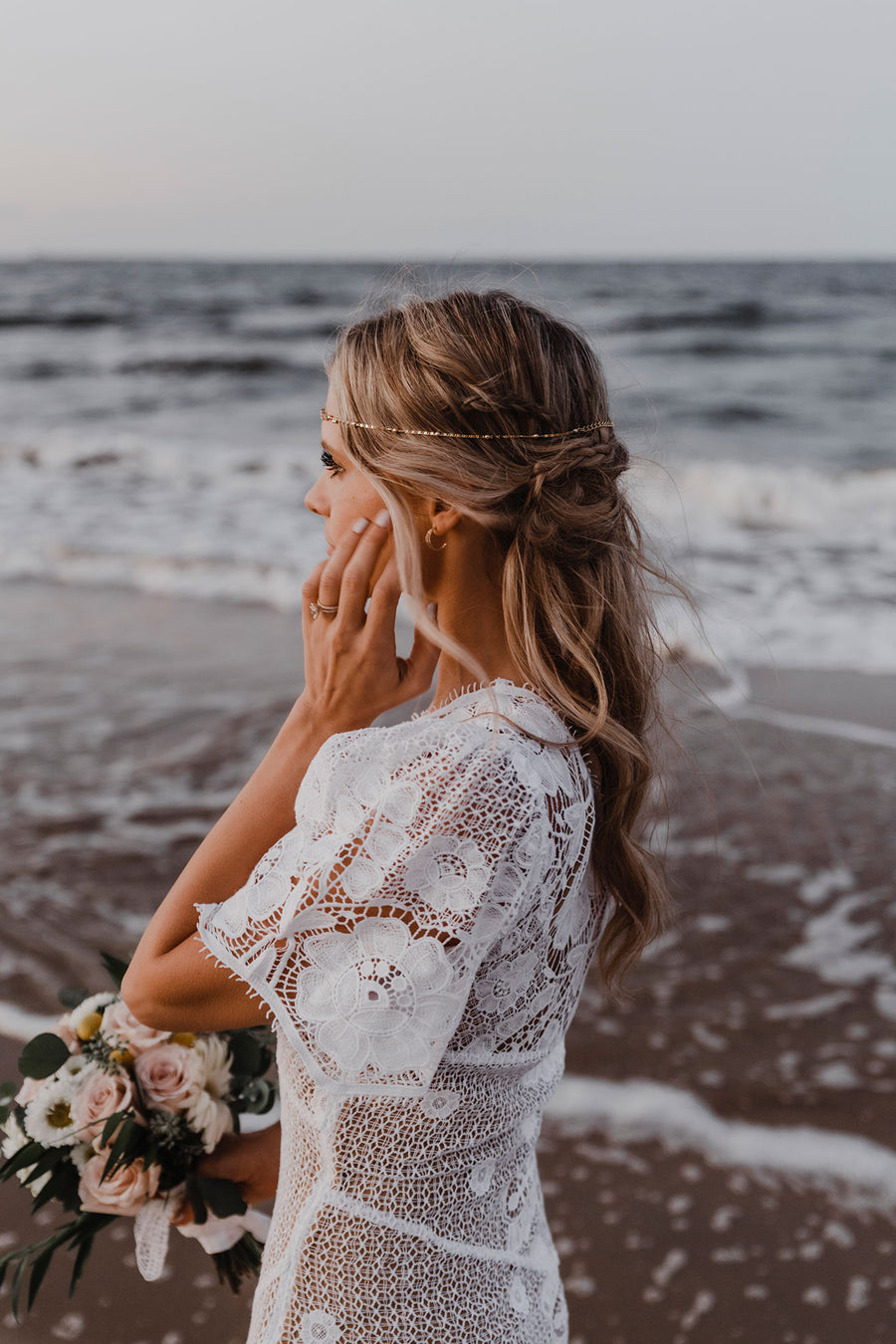 This is a side view photo of a bride wearing a floor length, one shoulder, maxi lace dress. She softly touches her face with her left hand. Her right hand holding a bouquet of flowers. She walks along the shoreline.