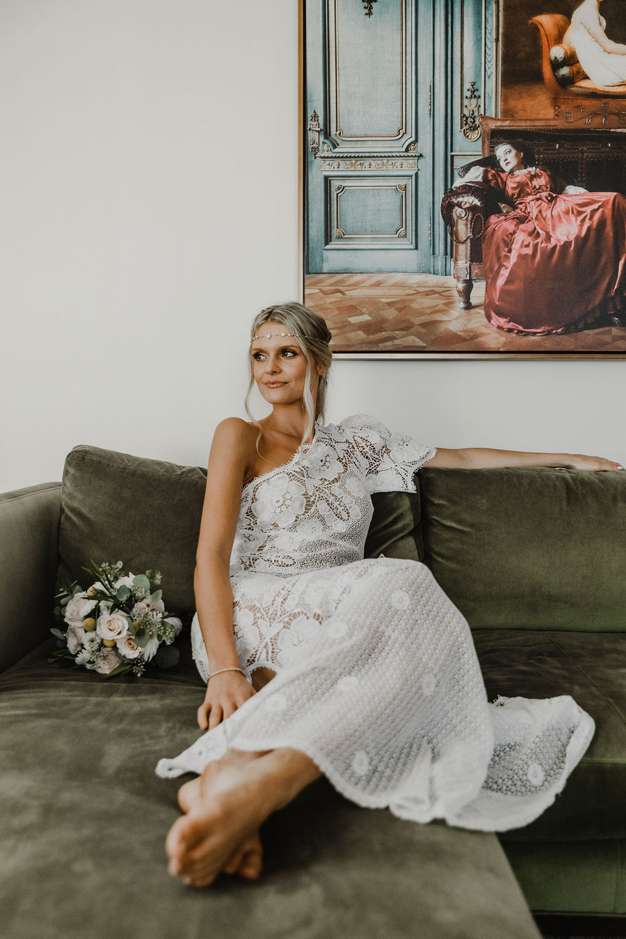 This is a photo of a woman wearing a floor length, one shoulder, maxi lace dress. There is a scalloped leg slit on the same side as the strapless top part of dress. The dress is lined to the leg slit. She sits on an olive green couch and is looking off into the distance.