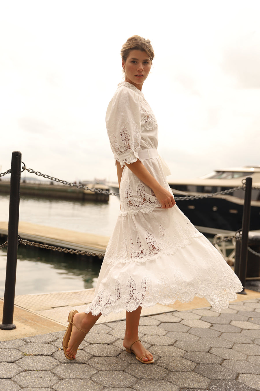 This is a photo of a woman wearing a long white linen and lace maxi dress with bow tie at the waist and puffy short sleeves. She spins in front of a body of water.