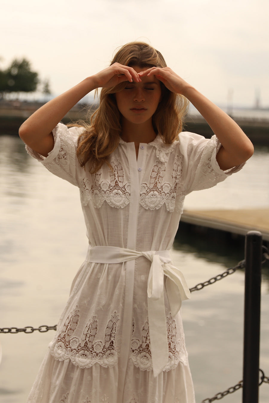This is a photo of a woman wearing a long white linen and lace maxi dress with bow tie at the waist and puffy short sleeves. She closes her eyes and has her hands on her head to shield the light.