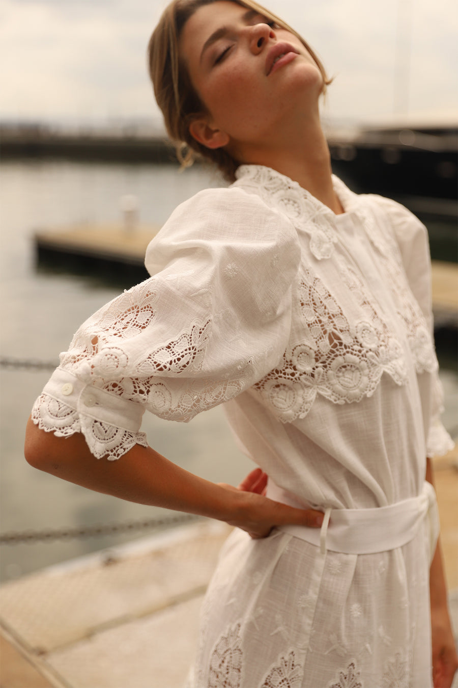 This is a photo of a woman wearing a long white linen and lace maxi dress with bow tie at the waist and puffy short sleeves. She looks up with her eyes closed and has one hand on her hip.