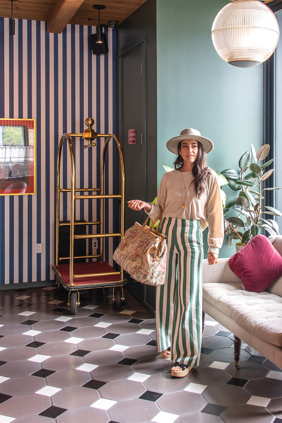 This is a photo of a woman wearing a white wide-brimmed hat and high waisted green and white striped wide leg pants with gold buttons. She is standing in a hotel lobby holding a floral tote.